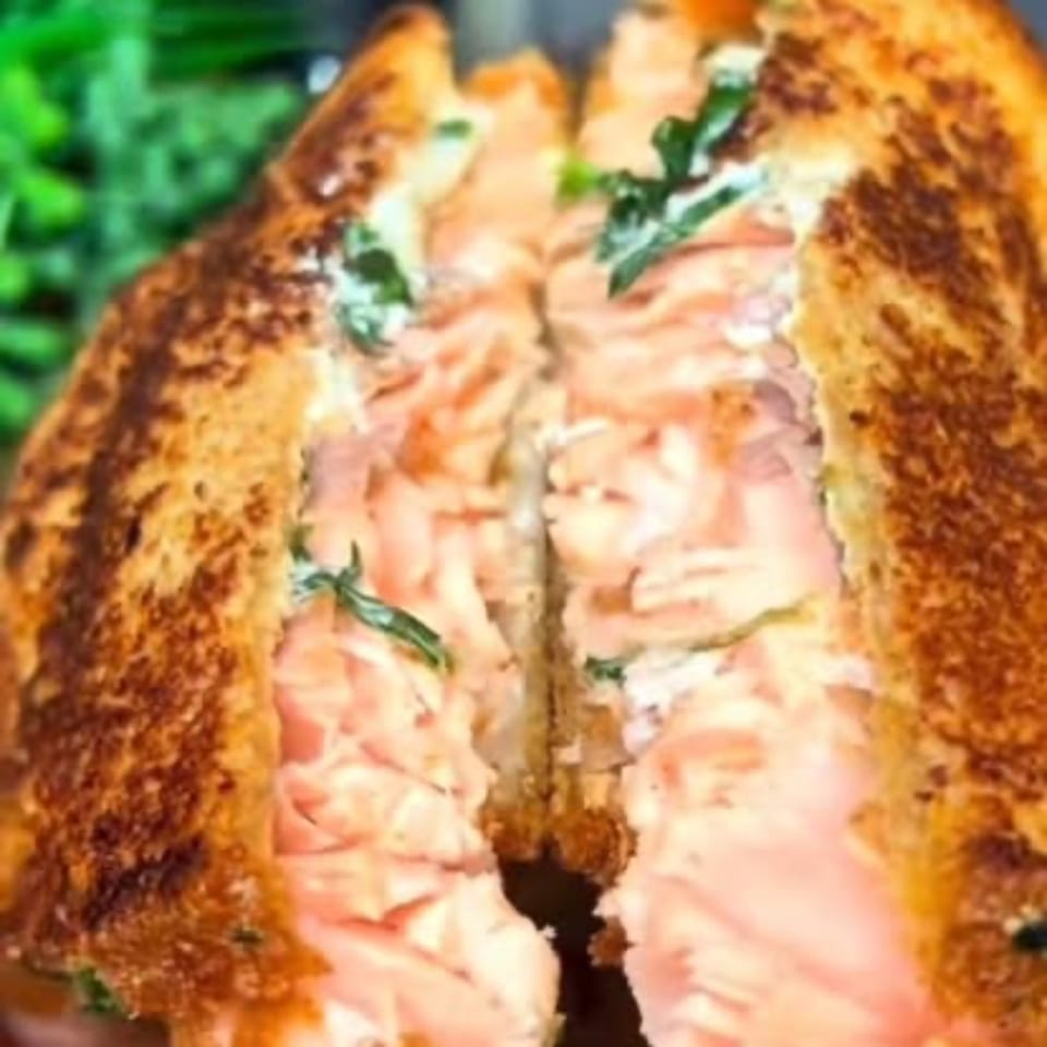 A grilled salmon sandwich, cut in half, revealing flaky pink fish and green herbs inside.