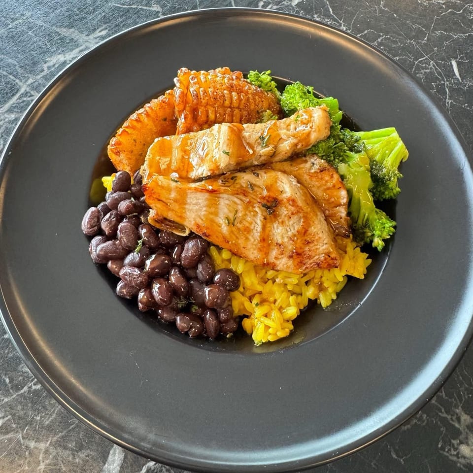 Grilled fish with yellow rice, black beans, corn on the cob, and broccoli on a black plate.