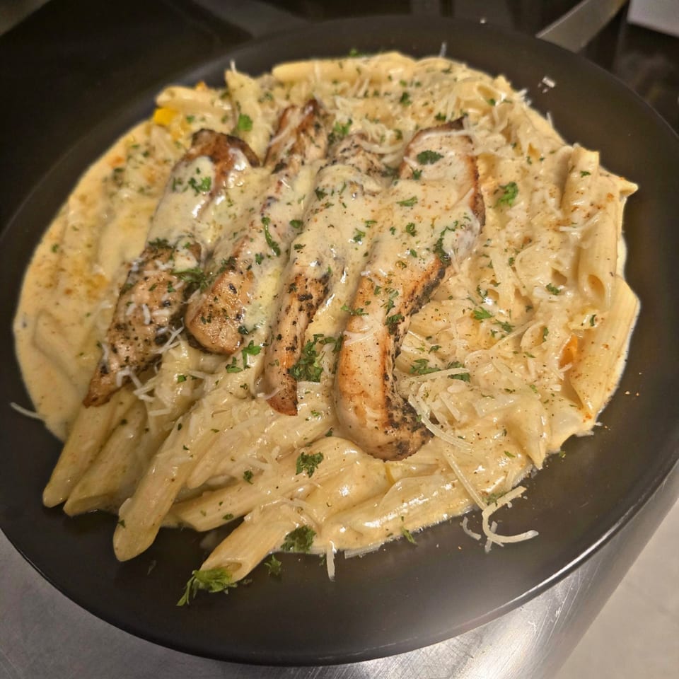 Creamy penne pasta with grilled chicken, parmesan cheese, and parsley in a black bowl.
